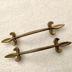 May include: Two antique brass drawer pulls with a fleur-de-lis design. Each pull features a long, slender bar with decorative ends. The brass has a warm, aged patina, suggesting vintage or antique hardware.