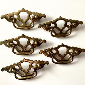 May include: Five antique brass drawer pulls with ornate, openwork designs. Each pull features a curved handle and decorative flourishes, perfect for vintage furniture restoration. The pulls are arranged on a white background.