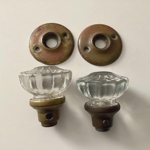 May include: Two antique glass door knobs with brass and bronze hardware. The clear glass knobs have a flower-like shape. Two matching bronze-colored backplates are above the knobs. The hardware is likely from the early 20th century.