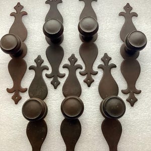 May include: A collection of antique-style, dark brown metal door pulls and knobs. Each piece features a rounded knob and an ornate, elongated backplate with decorative flourishes. The hardware is arranged in a symmetrical pattern.