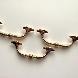 May include: Four gold-colored drawer pulls with a decorative design. Each pull has a curved handle with a central sphere and flared ends. The pulls are made of metal and have a shiny finish, suitable for furniture restoration or new builds.