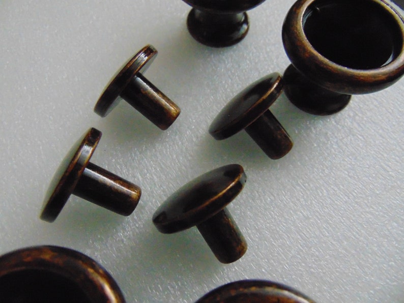 May include: A set of six antique bronze cabinet knobs. The knobs have a round base and a short, round stem. The knobs are in various positions on a white surface.