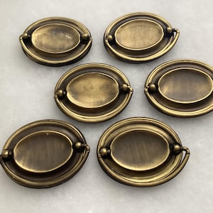 May include: A set of six oval-shaped brass drawer pulls, each featuring a central plate and a hinged drop handle. The metal has a muted, aged finish with visible variations in tone across the surface of each individual hardware piece.