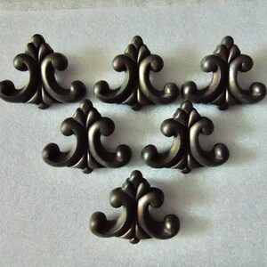 May include: Six bronze-colored decorative furniture appliques. Each applique features a symmetrical design with a central floral element and curved, scroll-like accents. The appliques are arranged on a light-colored surface.