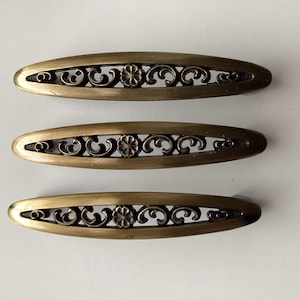 May include: Three antique brass-toned drawer pulls. Each oval pull features an openwork design with floral and scroll details. The pulls are arranged on a white surface, showcasing their vintage aesthetic. These decorative hardware pieces are ideal for furniture restoration projects.