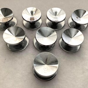 May include: A set of seven circular silver-toned metal buttons arranged in a triangular formation. Each button features a smooth, reflective, concave top surface that tapers toward the center, supported by a cylindrical base for attachment.
