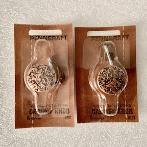 May include: Two copper-colored cabinet knobs with a textured design, packaged on brown cardboard backing. The knobs are round with a screw for attachment. The packaging reads "PENNCRAFT" and "CALICO KNOB".