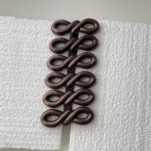 May include: A decorative, dark brown metal clasp featuring five interconnected infinity loop designs. The clasp is set against a textured white background, creating a contrast in color and texture. This clasp could be used for various craft projects.