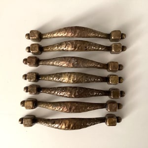 May include: Set of six antique bronze cabinet pulls. Each pull features a textured, curved handle with square end caps. The bronze has a warm, aged patina, adding to the vintage aesthetic. Ideal for furniture restoration or adding a rustic touch.