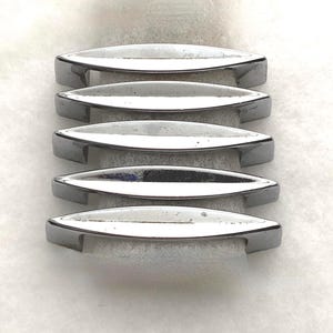 May include: A set of five silver-toned metal drawer pulls, each featuring a sleek, elongated, boat-like shape with pointed ends and a smooth, reflective finish. The handles are arranged in a vertical stack, showcasing their curved profile.