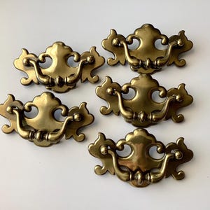 May include: Five antique brass drawer pulls with a decorative, ornate design. Each pull features a curved handle and a detailed backplate with a floral motif. The pulls are arranged on a white surface.