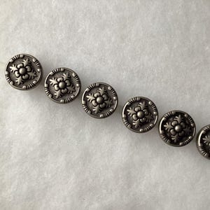 May include: A set of six round, metallic silver-toned buttons arranged in a diagonal line. Each button features a raised, symmetrical floral pattern in the center, surrounded by a textured, notched border along the outer edge of the circular face.