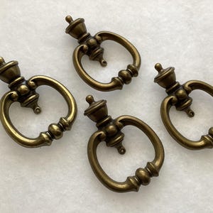 May include: A set of four antique brass-toned metal drawer pulls, each featuring a circular drop handle attached to a decorative mounting post with a small finial top and a rounded base plate with a single screw hole for attachment.