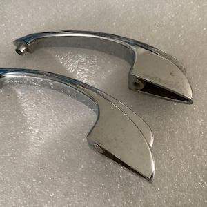May include: Two chrome-plated door handles. The handles have a curved design with a smooth, reflective surface. The handles are likely for a car or other vehicle. The handles are in good condition.