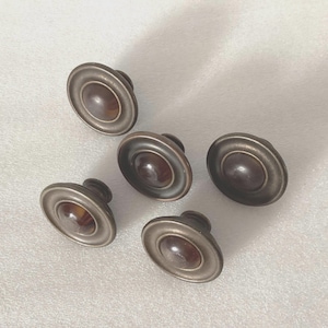 May include: Five antique-style cabinet knobs. Each knob features a dark brown, oval-shaped center surrounded by a silver-colored metal rim. The knobs are arranged on a white surface.