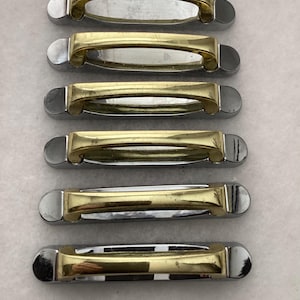 May include: A set of six horizontal drawer pulls, each featuring a polished gold-tone metal handle mounted on a rectangular chrome-finished base. The hardware displays a two-tone metallic design with rounded ends and a central open grip area.