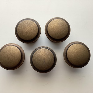 May include: Five antique brass cabinet knobs. Each knob has a round, slightly domed center and a circular base. The knobs have a weathered, aged appearance, with variations in color and texture. These knobs are suitable for furniture restoration or DIY projects.