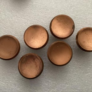 May include: Six copper-colored, round cabinet knobs with a slightly domed shape are arranged on a light-colored surface. The knobs have a brushed finish, and some show minor wear.