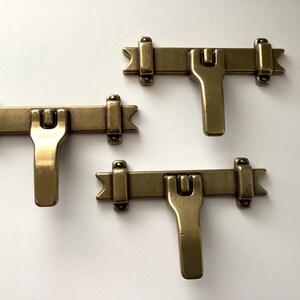 May include: Three antique brass latches. Each latch has a rectangular bar with decorative ends and a central U-shaped handle. The latches are arranged on a white surface, showcasing their aged brass finish and classic design.
