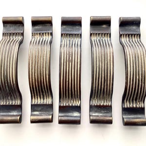 May include: Five antique bronze cabinet pulls. Each pull has a curved, ribbed design with a dark bronze finish. The pulls are arranged in a row against a white background. These decorative hardware pieces are suitable for furniture restoration or new builds.