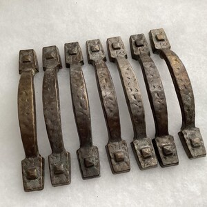 May include: A set of seven metal drawer pulls with a dark, hammered bronze finish. Each handle features a curved center grip and rectangular mounting plates at both ends, each accented with a small, raised square detail in the center.