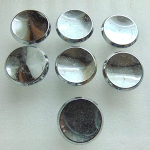 May include: A set of seven circular, metallic buttons with a polished, reflective silver finish. Each button features a smooth, slightly concave surface and a raised, rounded edge, presenting a uniform appearance across the entire collection.
