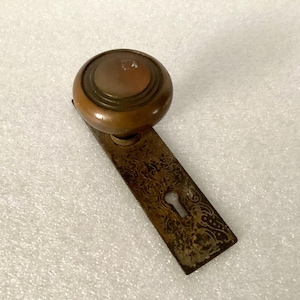 May include: Antique bronze doorknob with a round knob and a rectangular backplate. The backplate features an ornate, etched design and a keyhole. The doorknob is a warm, copper color, and the backplate has a weathered appearance.