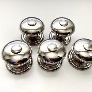 May include: Six shiny, silver-toned cabinet knobs with a classic design. Each knob features a rounded top with a smaller, circular accent and a cylindrical base. The knobs are arranged on a white surface, reflecting light.