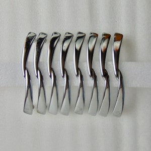 May include: A set of eight shiny, chrome-finished golf clubs, arranged in a row. Each club has a curved, elongated head and a sleek, reflective surface. The clubs are set against a plain white background.