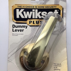 May include: A Kwikset Plus dummy lever door handle in a clear plastic package. The handle is gold-colored and has a curved lever design. The packaging includes the text "THE #1 CHOICE IN HOME SECURITY" and "Dummy Lever".