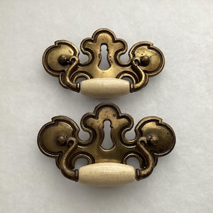 May include: Two antique brass drawer pulls with a keyhole design and cream-colored ceramic handles. The pulls have an ornate, vintage aesthetic, perfect for adding a touch of elegance to furniture.
