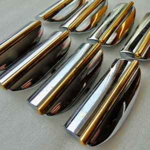 May include: A collection of chrome and gold-toned drawer pulls. Each pull has a curved, polished chrome body with a central gold-colored accent. The pulls are arranged in a group, showcasing their reflective surfaces and design.