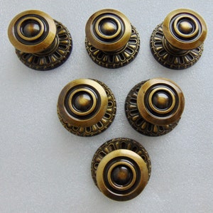 May include: Six antique bronze-colored cabinet knobs. Each knob features a central circular design surrounded by concentric rings and a decorative, scalloped base. The knobs are arranged on a light-colored surface.