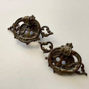 May include: Two antique bronze drawer pulls with ornate detailing. Each pull features a circular handle, decorative backplate, and intricate floral accents. The hardware is aged with a patina finish.