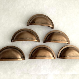 May include: A set of six semi-circular cup drawer pulls in a weathered bronze finish. Each pull features a rounded, arched handle design with a flat bottom edge and a smooth, metallic surface with subtle dark patina accents throughout the metal.