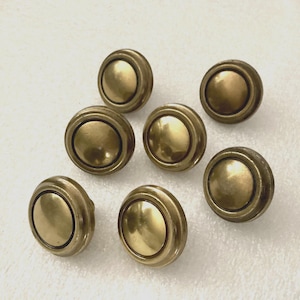 May include: A collection of seven antique brass cabinet knobs. Each knob features a circular, slightly domed center surrounded by a ridged border. The knobs have a warm, aged brass finish, suggesting vintage hardware.
