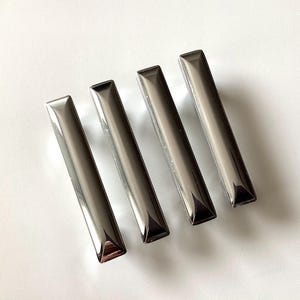 May include: Four rectangular, metallic silver cabinet pulls are arranged on a white surface. Each pull has a rectangular shape with a slightly beveled top and bottom. The pulls are evenly spaced and reflect the light, creating a shiny appearance.