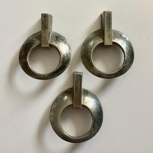 May include: Three silver-toned metal drawer pulls with a geometric design. Each pull has a circular ring with a vertical bar in the center.