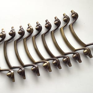 May include: A collection of nine antique brass drawer pulls. Each pull has a curved design with a decorative finial and a rounded knob. The pulls are arranged in a row against a white background.