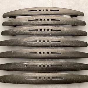 May include: A set of seven dark metallic drawer pulls arranged in a vertical stack. Each handle features a curved, elongated shape with a central decorative cutout consisting of three small squares flanked by two horizontal rectangular slots.