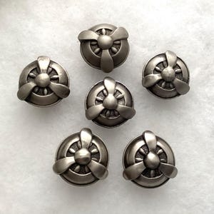 May include: A set of six circular metal buttons, each featuring a raised three-blade propeller design in a matte silver finish. The center of each button has a smooth dome, with textured detailing visible behind the propeller blades.
