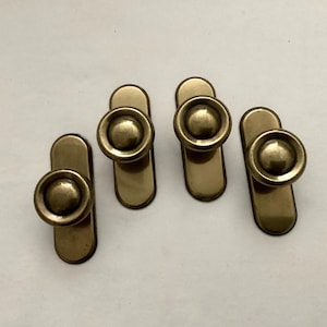 May include: Four antique brass cabinet pulls. Each pull features a round knob mounted on a rectangular backplate. The brass has a warm, aged patina, suggesting vintage hardware. These pulls are suitable for drawers or cabinet doors.