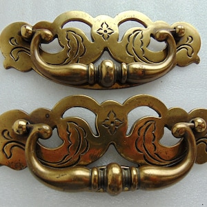 May include: Two antique brass drawer pulls. Each pull features a curved handle and decorative backplate with floral and starburst designs. The brass has a warm, aged patina, suggesting vintage or retro style. The pulls are likely for furniture restoration.