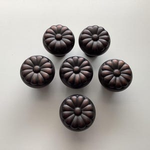 May include: Six bronze-colored cabinet knobs with a floral design. Each knob has a rounded shape with a detailed, petal-like pattern radiating from the center. The knobs are arranged in a circular pattern.