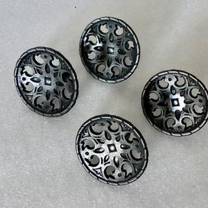 May include: Four silver-toned, oval-shaped buttons with intricate openwork designs. Each button features a central square and surrounding floral and scroll patterns. The buttons are arranged on a light-colored surface.