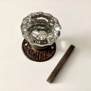May include: A clear, faceted glass doorknob with a decorative, ornate bronze base and a rusty metal key. The doorknob is a vintage style, likely for a door. The key is rectangular with a series of teeth.