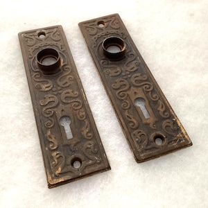 May include: Two rectangular bronze-toned metal door plates featuring an embossed scrollwork pattern. Each plate includes a circular opening for a door knob at the top and a keyhole opening at the bottom, with small screw holes at both ends.