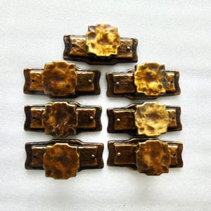 May include: Set of six antique-style cabinet pulls. Each pull features a rectangular base with a central, textured, golden-bronze accent. The pulls are made of a dark bronze-colored metal and are arranged in a grid pattern.