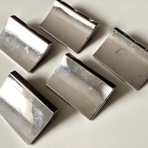 May include: Six polished, rectangular metal drawer pulls with a curved top edge. The pulls are silver in color and arranged on a light-colored surface. The pulls have a reflective surface.