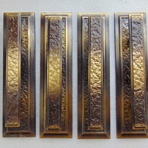May include: Six rectangular bronze-toned metal door pulls. Each pull features a textured, central panel framed by a smooth, stepped border. The metal has a gradient of colors, from dark bronze to gold.